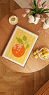 una mesa de madera con una imagen enmarcada de una naranja, un cuenco de limones, un jarrón con tulipanes rosas y una vela.
