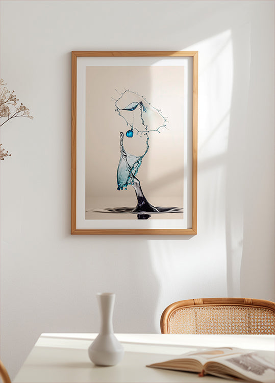 una obra de arte enmarcada de una gota de agua suspendida en el aire, con una combinación de colores azul y blanco. La obra de arte se exhibe en una pared blanca sobre una mesa con un jarrón blanco y un libro.