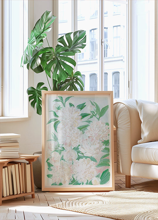 una gran impresión enmarcada de flores blancas con hojas verdes, colocada sobre un piso de madera en una habitación amplia y luminosa con grandes ventanales.